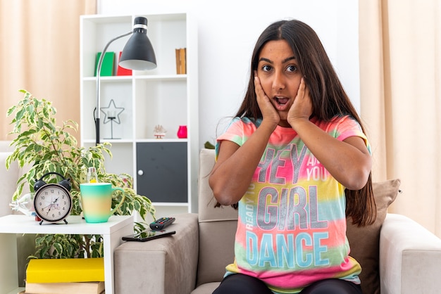 Menina com roupas casuais parecendo espantada e surpresa, sentada na cadeira na sala de estar iluminada