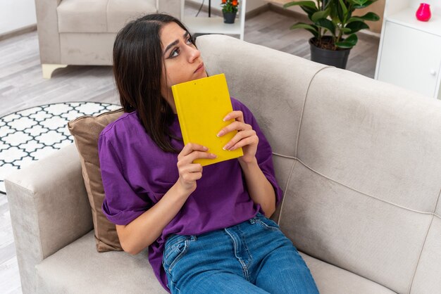 Menina com roupas casuais segurando um livro, olhando para o lado, perplexa, sentada em um sofá em uma sala iluminada