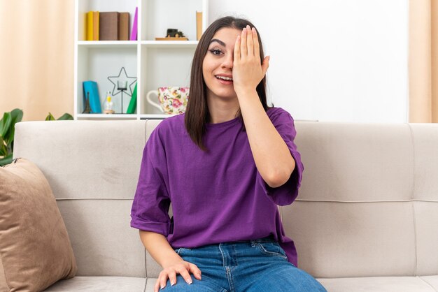 Menina com roupas casuais sorrindo alegremente cobrindo um olho com a mão sentada em um sofá na sala de estar iluminada