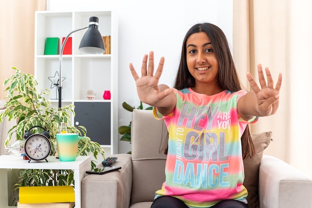 Menina com roupas casuais, sorrindo alegremente, mostrando o número nove com os dedos sentados na cadeira na sala iluminada