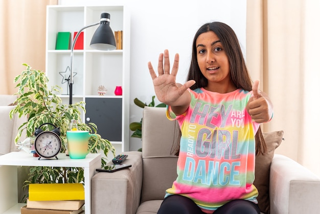 Menina com roupas casuais sorrindo confiante mostrando o número cinco com a palma da mão aparecendo os polegares para cima, sentada na cadeira na sala iluminada