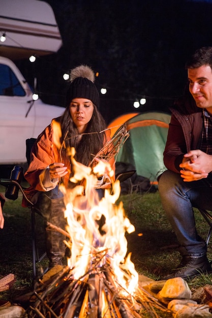 Menina com seus amigos relaxando juntos no acampamento ao redor da fogueira nas montanhas. Carrinha de campista retro.