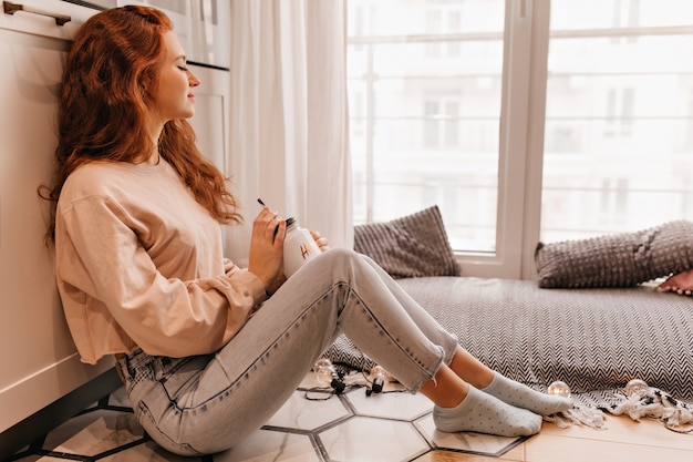 Foto grátis menina com sono em jeans, bebendo vinho quente em dia frio. foto interna de jovem encaracolado posando com uma xícara de chá.