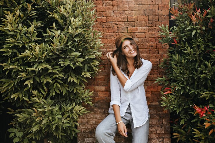 Foto grátis menina com sorrisos encantadores de covinhas. mulher de camisa da moda resort e calças posando relaxado em um espaço de tijolos com arbustos.
