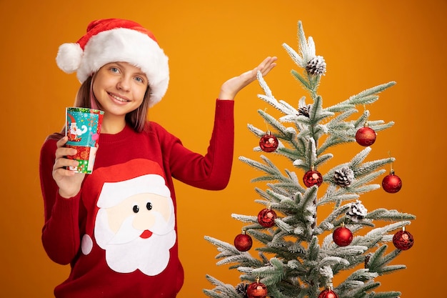 Menina com suéter de Natal e chapéu de Papai Noel segurando um copo de papel colorido, apresentando-se com o braço da mão sorrindo ao lado de uma árvore de Natal sobre fundo laranja