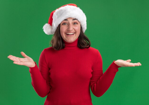 Menina com suéter vermelho e chapéu de Papai Noel olhando para a câmera com uma cara feliz abrindo os braços para os lados em pé sobre um fundo verde