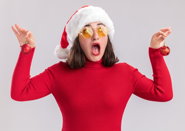 Menina com um suéter vermelho e chapéu de Papai Noel usando óculos segurando bolas de natal, olhando de lado surpresa em pé sobre um fundo branco