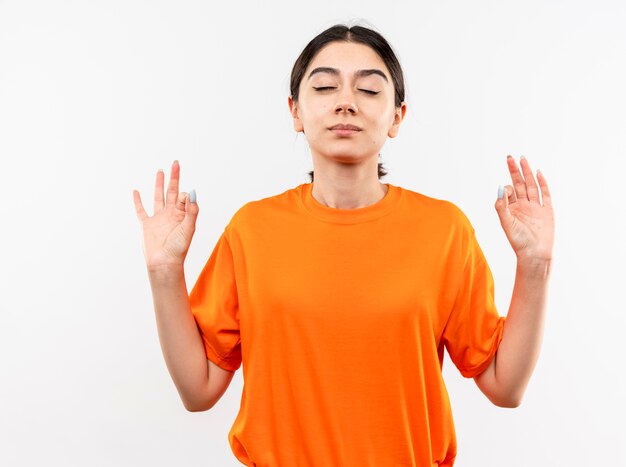 Menina com uma camiseta laranja relaxando com os olhos fechados fazendo um gesto de meditação com os dedos em pé sobre uma parede branca