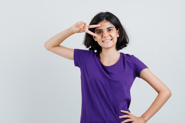 Menina com uma camiseta mostrando o sinal V perto do olho e parecendo alegre,