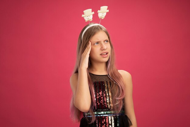 Menina com vestido de festa brilhante e bandana engraçada olhando de lado confusa, conceito de feriado de celebração de ano novo em pé sobre fundo rosa