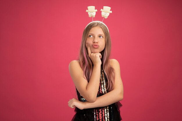 Menina com vestido de festa brilhante e bandana engraçada olhando de lado confusa, conceito de feriado de celebração de ano novo em pé sobre fundo rosa