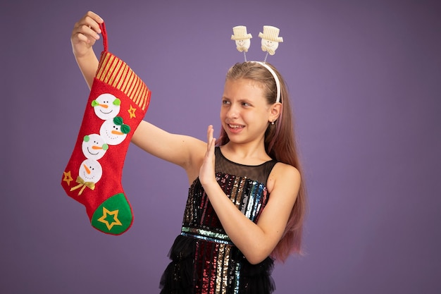 Menina com vestido de festa glitter e bandana engraçada segurando uma meia de natal olhando para ela descontente, segurando a mão de pé sobre o fundo roxo