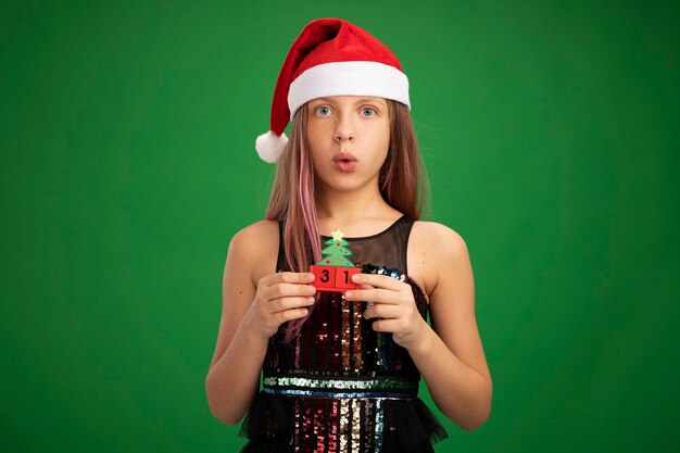 Menina com vestido de festa glitter e chapéu de Papai Noel segurando cubos de brinquedo com data de ano novo olhando para a câmera surpresa em pé sobre fundo verde