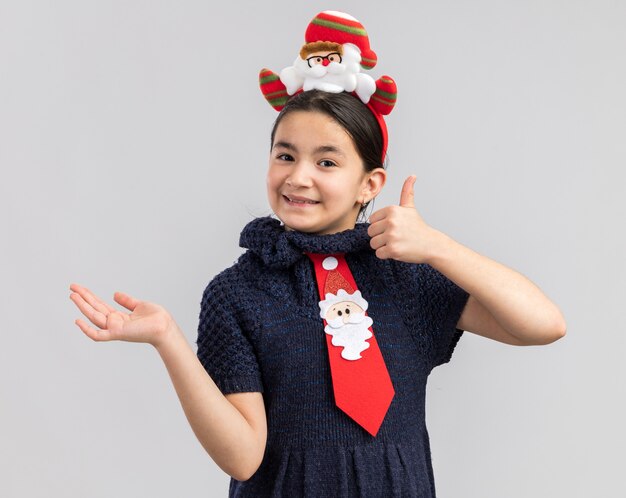 Menina com vestido de malha, gravata vermelha com aro de natal engraçado na cabeça, sorrindo, mostrando os polegares para cima, apresentando o espaço da cópia com o braço da mão