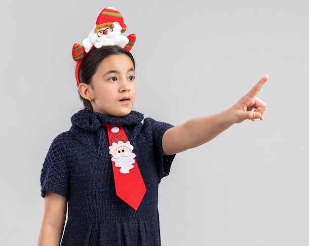 Menina com vestido de malha usando gravata vermelha com aro de natal engraçado na cabeça apontando com o dedo indicador para algo preocupado