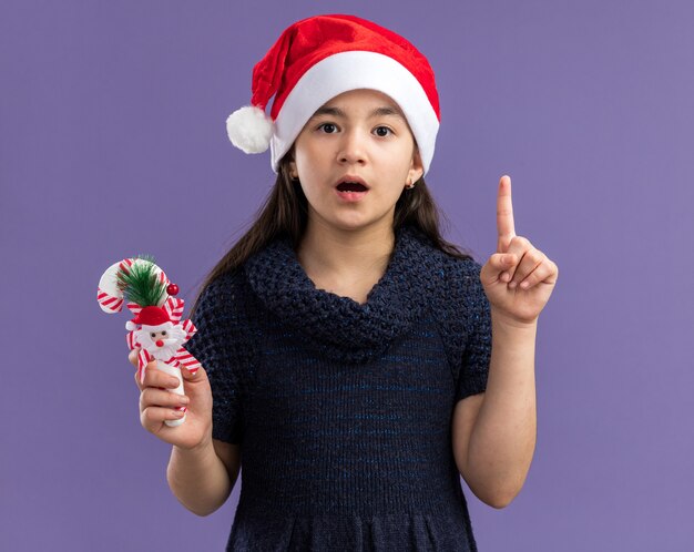 Menina com vestido de tricô com chapéu de Papai Noel segurando bengala de Natal surpresa mostrando o dedo indicador de pé sobre a parede roxa