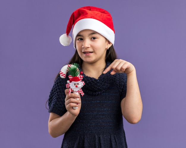 Menina com vestido de tricô com chapéu de Papai Noel segurando o bastão de doces de natal apontando com o dedo indicador para ele com um sorriso no rosto em pé sobre a parede roxa