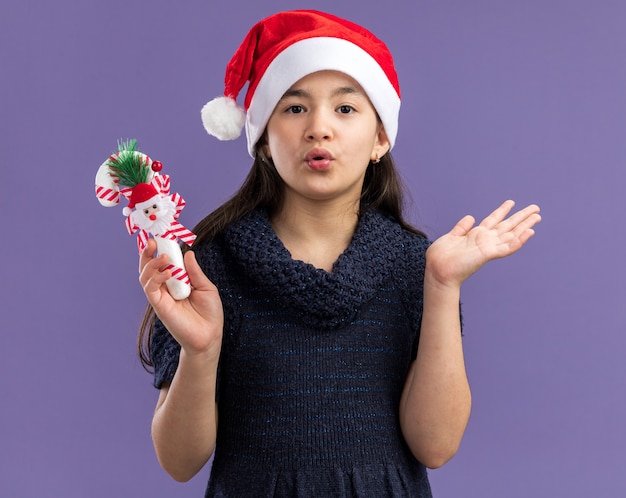 Menina com vestido de tricô com chapéu de papai noel segurando uma bengala de doces de natal surpresa com os braços levantados em pé sobre a parede roxa