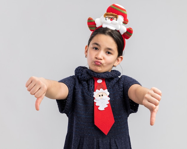 Menina com vestido de tricô usando gravata vermelha com aro de natal engraçado na cabeça parecendo descontente mostrando os polegares para baixo