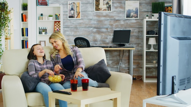 Menina comendo batatinhas enquanto assiste tv com a mãe dela sentada no sofá.