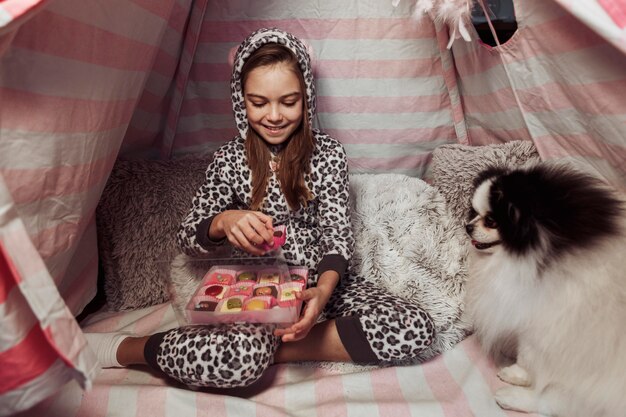 Menina comendo doces em uma barraca dentro de casa e um cachorro