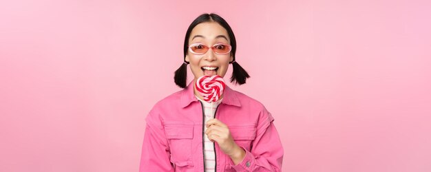 Menina coreana estilosa lambendo pirulito comendo doces e sorrindo de pé em óculos de sol contra fundo rosa