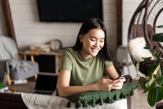 Menina coreana sorridente, conversando no celular, sentada em casa na sala de estar com smartphone e risos ...