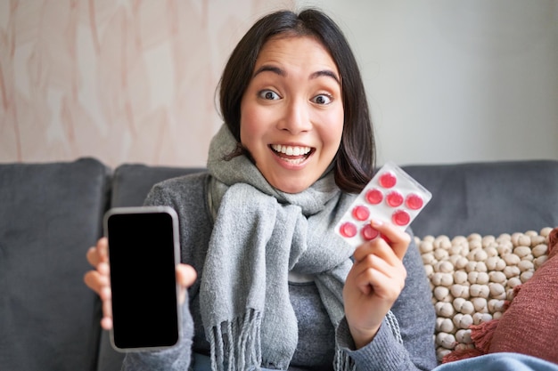 Menina coreana sorridente mostra medicação de tela de smartphone nas mãos sentindo-se doente e ficando em casa usi