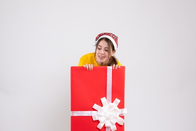 menina curiosa com chapéu de Papai Noel olhando algo atrás de um grande presente de Natal em branco