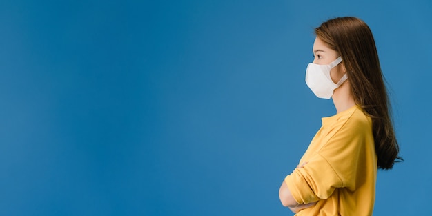 Menina da ásia jovem usando máscara médica com vestido de pano casual e olhar para o espaço em branco isolado sobre fundo azul. distanciamento social, quarentena para o vírus corona. fundo do banner panorâmico.