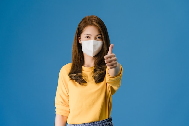 Menina da Ásia jovem usando máscara médica aparecendo polegar com vestido de pano casual e olhar para a câmera isolada sobre fundo azul. Auto-isolamento, distanciamento social, quarentena para vírus corona.