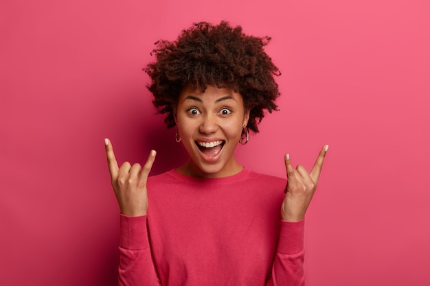 Menina de cabelo encaracolado positiva faz gestos de chifre, gosta de música rock n roll, ri de alegria e usa blusão casual