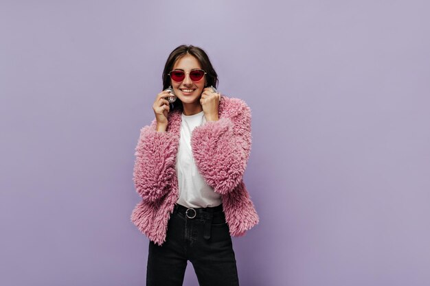 Menina de cabelo morena com óculos de sol vermelhos em calça preta de camiseta branca com cinto e suéter fofo rosa sorrindo e segurando bolas de discoteca