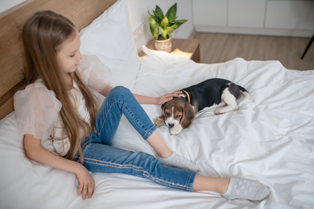 Menina de cabelos compridos sentada na cama dando tapinhas na cabeça de seu lindo amigo de quatro patas