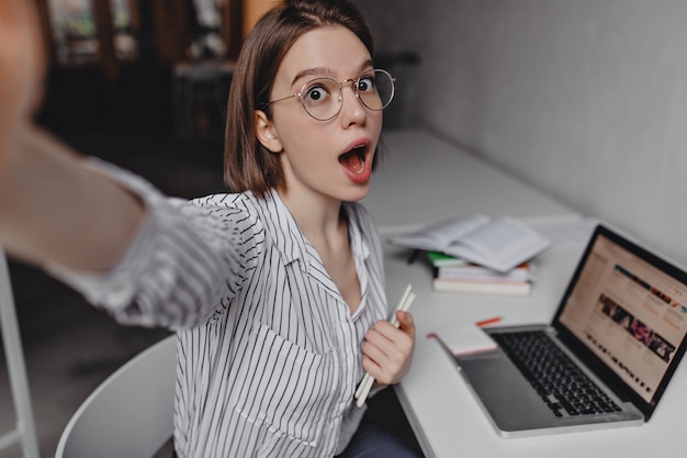 Foto grátis menina de cabelos curtos de olhos castanhos na blusa branca, segurando o caderno e tomando selfie no fundo do laptop e material de escritório.