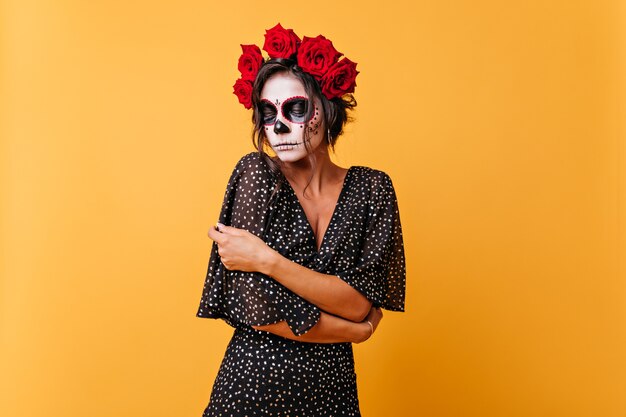 Menina de cabelos escuros envergonhada com flores na cabeça posando no crânio de Calavera tradicional. Retrato de uma modelo mexicana olhando para o chão