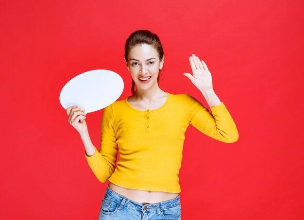 Menina de camisa amarela segurando um quadro oval de informações e cumprimentando alguém