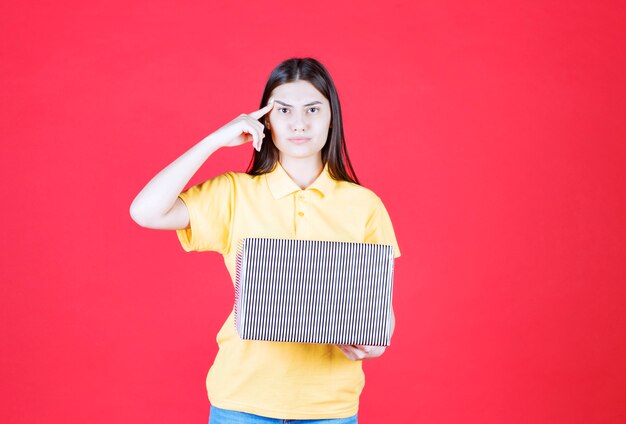 Menina de camisa amarela segurando uma caixa de presente prata e parece confusa e pensativa