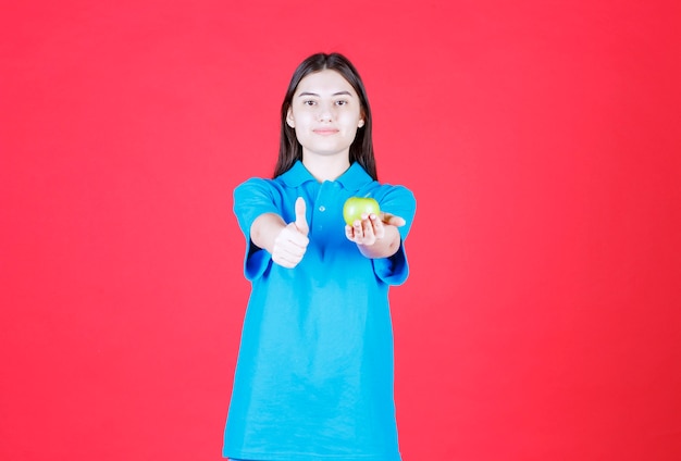 Menina de camisa azul segurando uma maçã verde e mostrando sinal positivo