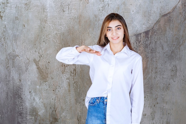 Menina de camisa branca em pé em uma parede de concreto e mostrando a altura de um objeto.
