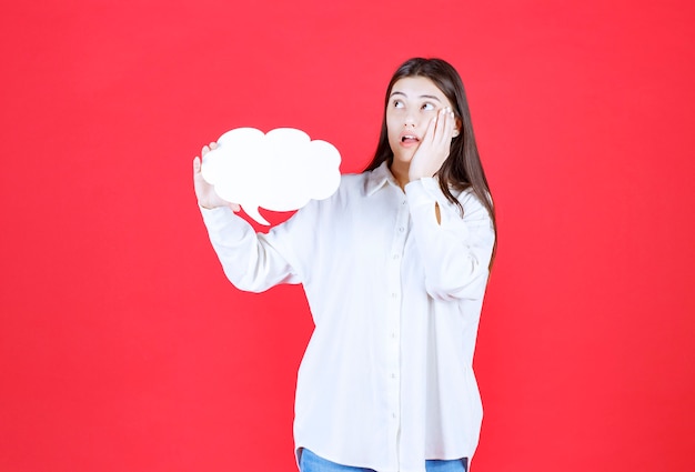 Menina de camisa branca segurando um quadro de informações em forma de nuvem e parece emocionada e apavorada