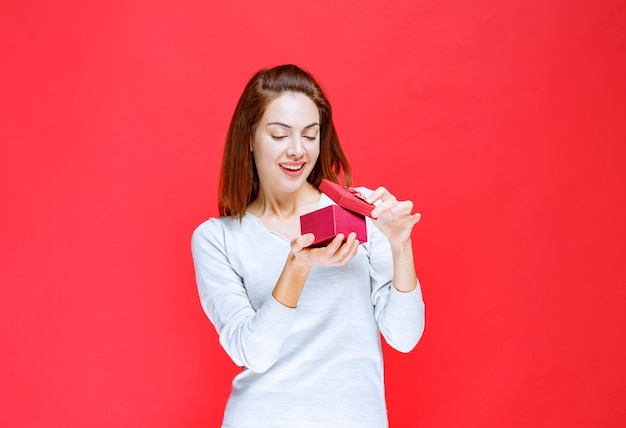 Menina de camisa branca segurando uma pequena caixa de presente vermelha, abrindo-a e ficando surpresa.