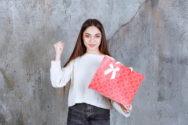 Menina de camisa branca segurando uma sacola de compras vermelha e mostrando sinal de mão positiva.