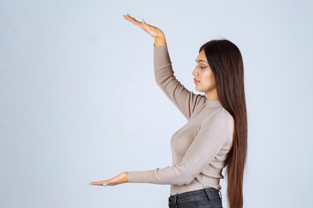 Menina de camisa cinza, mostrando a altura de um objeto.