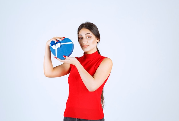 Menina de camisa vermelha, apresentando sua caixa de presente de forma de coração azul.