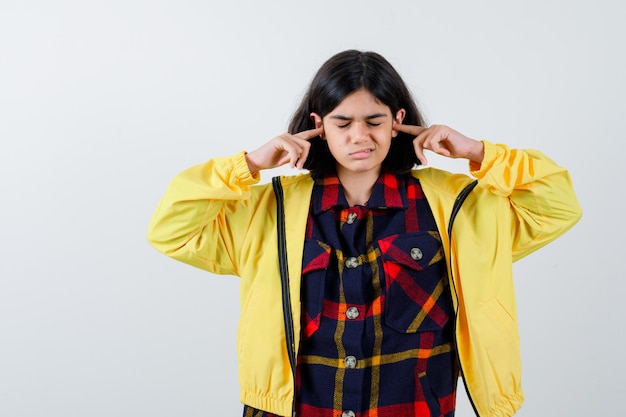 Foto grátis menina de camisa xadrez, jaqueta, tampando as orelhas com os dedos e parecendo irritada, vista frontal.