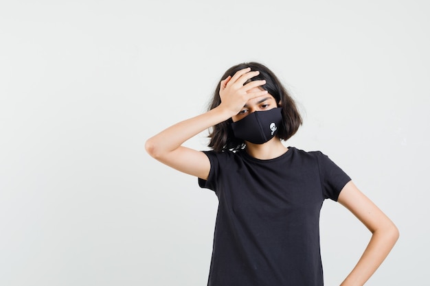 Menina de camiseta preta, máscara, segurando a mão na cabeça e parecendo exausta, vista frontal.