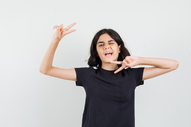 Menina de camiseta preta, mostrando o gesto de vitória, piscando os olhos, vista frontal.