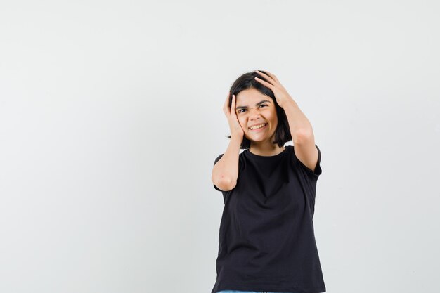 Menina de camiseta preta, segurando a cabeça com as mãos e olhando feliz, vista frontal.