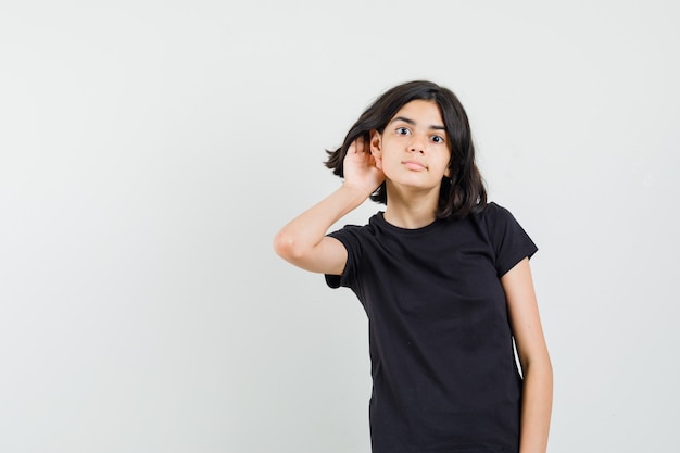 Menina de camiseta preta, segurando a mão atrás da orelha e olhando curiosa, vista frontal.